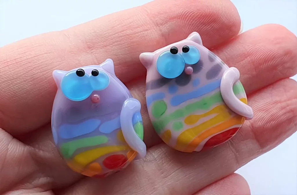 Two lampwork glass beads in lavender, resembling cats with kooky eyes and rainbow streaks.