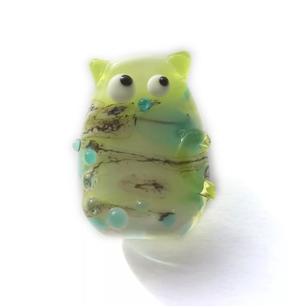 Lampwork glass bead of a stylized cat, with kooky eyes looking sideways. The bead has glass dots and a semi-transparent layer, making the inner colors pop. It's mainly lime green and and cyan, with some silicate layers intersperesed.