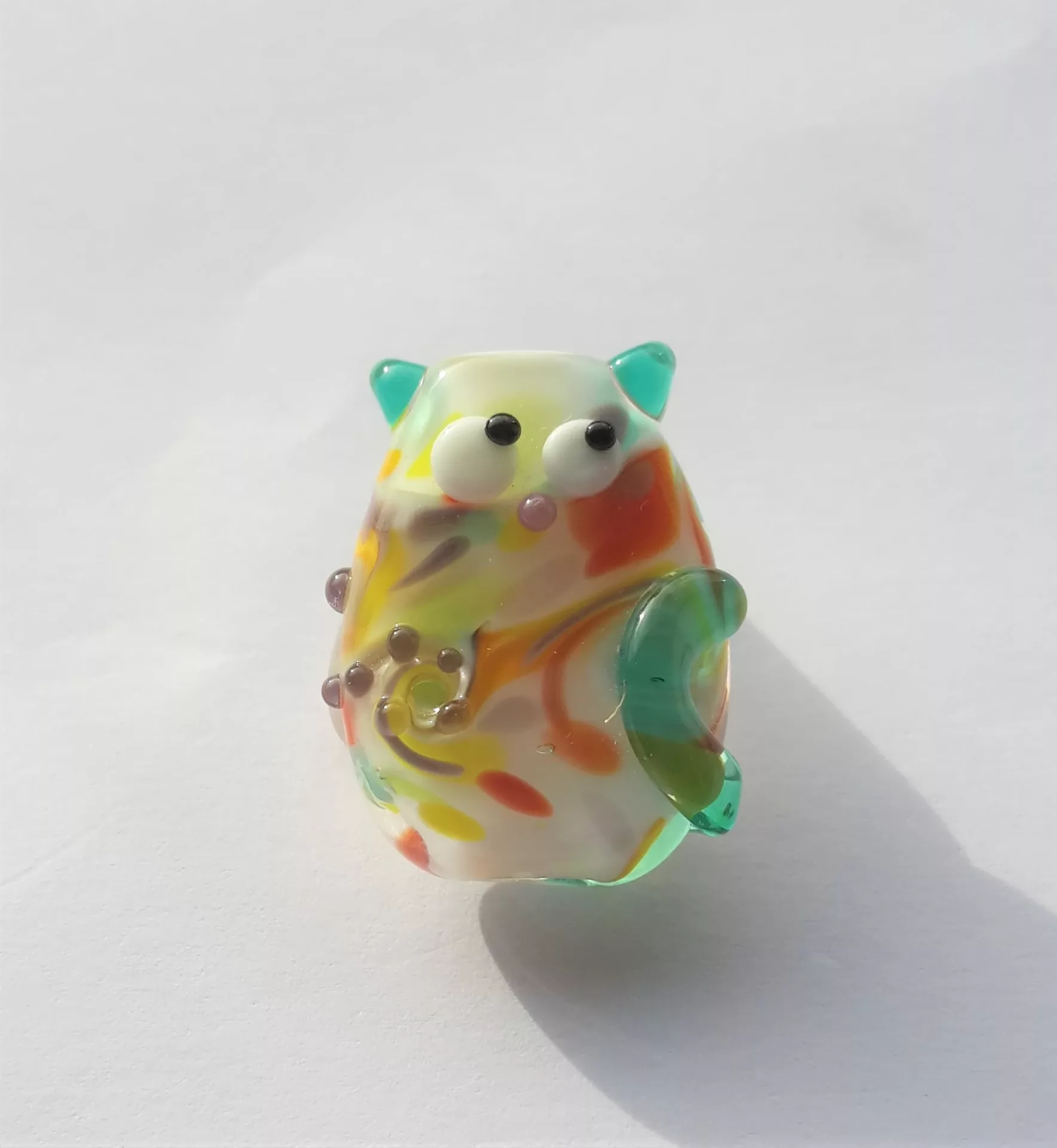 Lampwork glass bead resembling cat with kooky eyes looking sideways. Pearls and dots are being twisted by a swirl into the body of the cat.