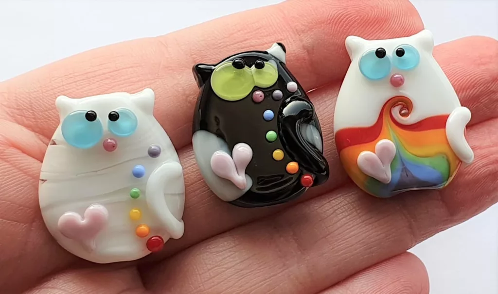 Three lampwork glass beads resembling kooky cats. They're decorated with pink hearts, two of them have rainbow dots and the last a rainbow swirl.