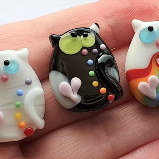 Three lampwork glass beads resembling kooky cats. They're decorated with pink hearts, two of them have rainbow dots and the last a rainbow swirl.